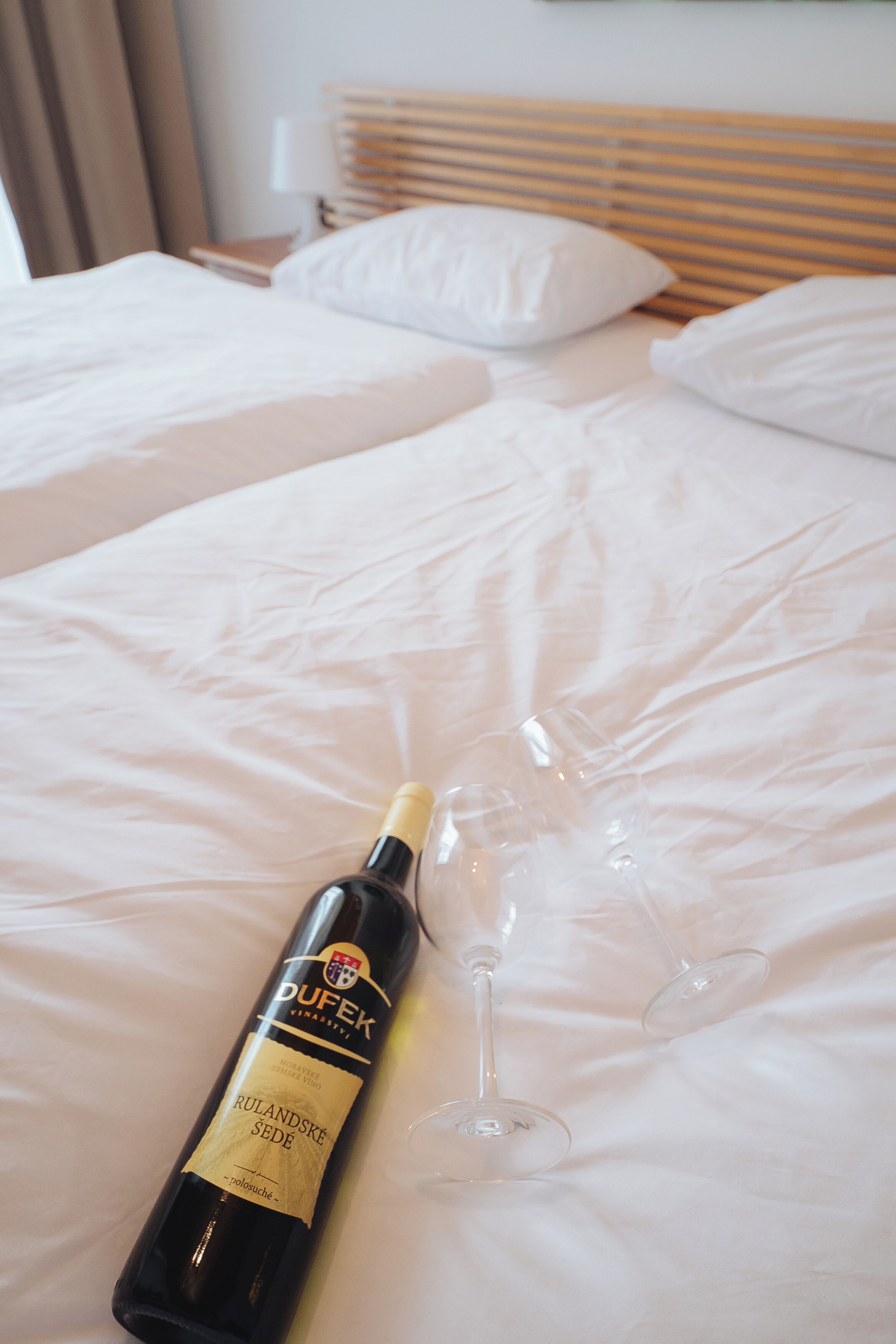 A bottle of Rulandske Sede wine and two wine glasses on a bed.