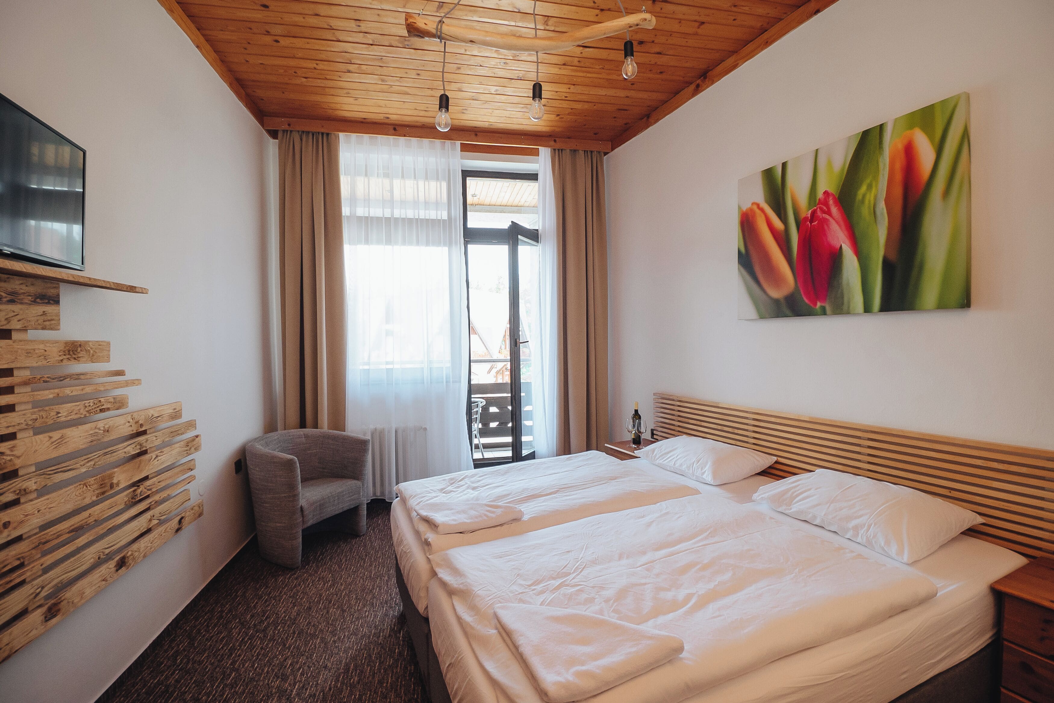 A hotel room with a wooden ceiling, a bed with white linens, a gray armchair, a painting of tulips, and a TV mounted on the wall.