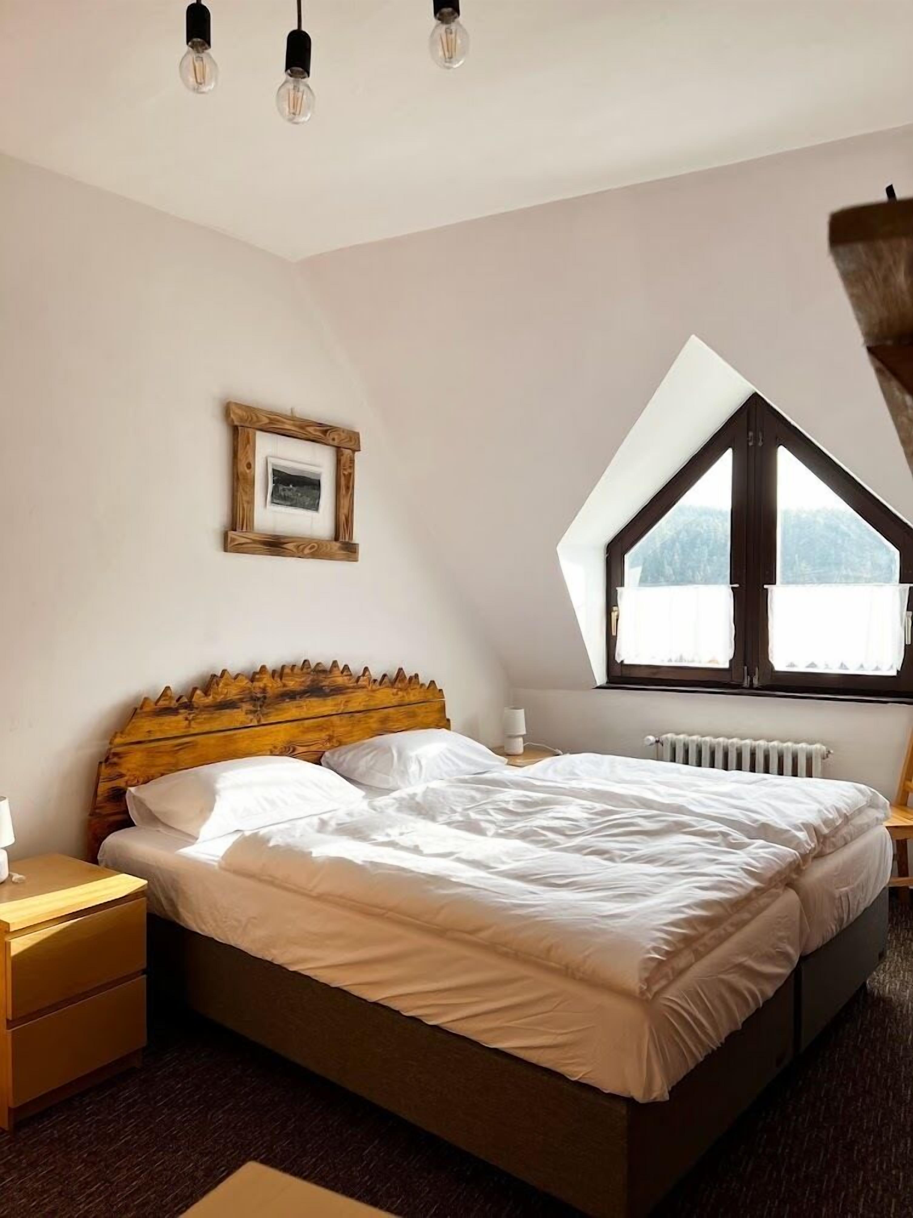 A bedroom with a sloped ceiling, a wooden headboard, a bed with white linens, a nightstand, a bedside table, a framed picture, and a window with a view.