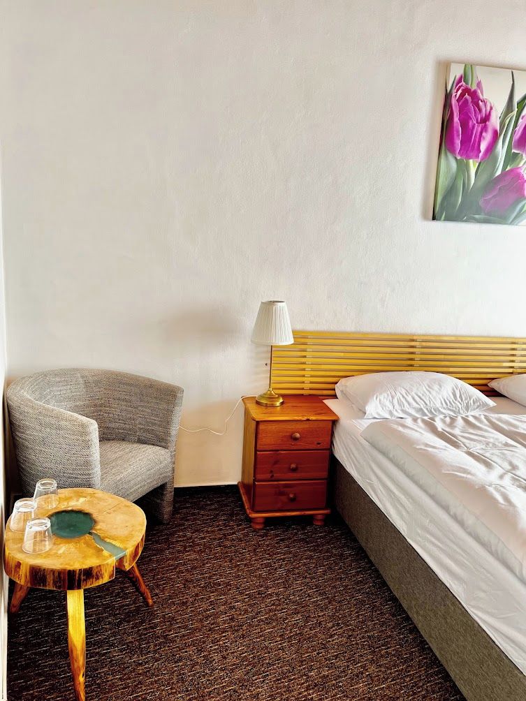 A hotel room with a bed, a chair, a small wooden table, and a lamp.