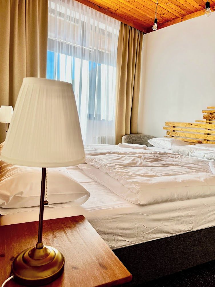 Una habitación de hotel con cama, mesita de noche con lámpara, cabecero de madera y ventana con cortinas.