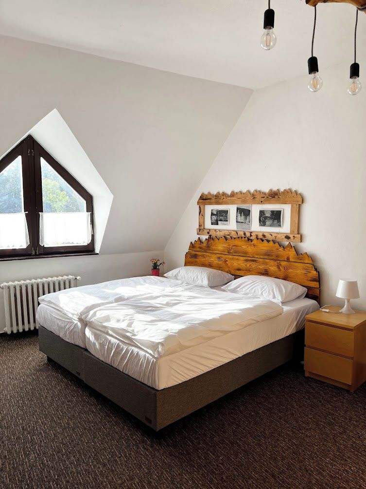 A bedroom with a sloped ceiling, a bed with white linens, a wooden headboard, a bedside table with a lamp, and a window with a view.