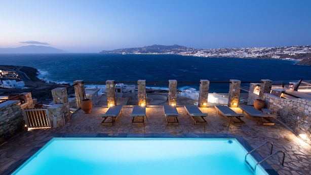 Zona de piscina con muros de piedra, sillas de descanso y vistas al mar y a la ciudad distante.