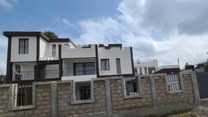 A modern multi-story residential building with a white exterior and dark trim, featuring balconies and a brick wall in the foreground. - Astrahill Villa,  A Royal Gateway near Ngong Hill. Nairobi (Kajiado County)