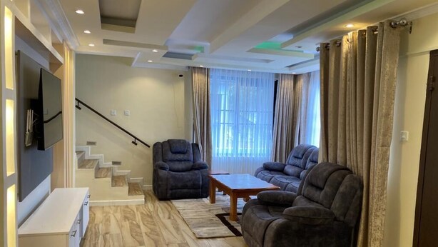 A modern living room with a sofa set, a coffee table, and a TV mounted on the wall. There is a staircase leading to another level. - Astrahill Villa,  A Royal Gateway near Ngong Hill. Nairobi (Kajiado County)
