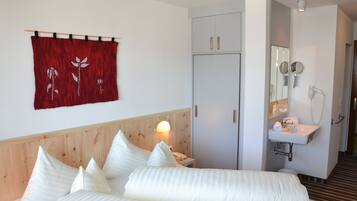 A hotel room with a bed, a wooden headboard, a red curtain with floral embroidery, a white bedside table with a lamp, a white sink with a mirror, and a door leading to another room.