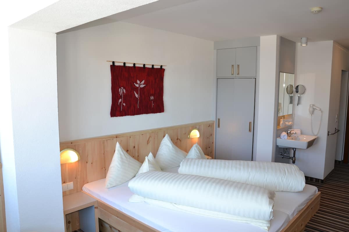 Une chambre d’hôtel comprenant un lit, des lampes de chevet, une tête de lit en bois, une décoration murale rouge, un miroir et une salle de bain avec un lavabo et un miroir.