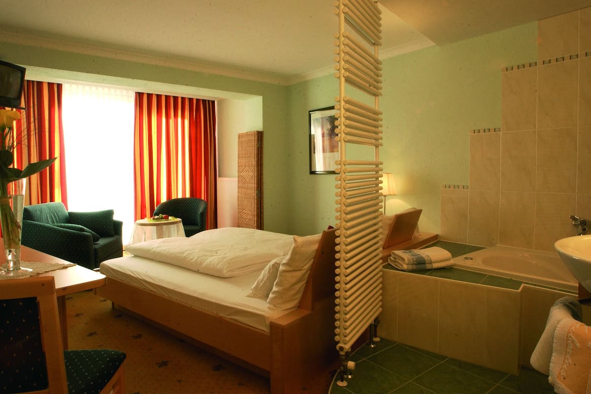 Une chambre d’hôtel comprenant un lit, une chaise, un bureau avec une lampe, une salle de bain avec une baignoire et un lavabo, et une fenêtre avec des rideaux.