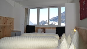 A hotel room with a large bed, a wooden headboard, a small desk, and a view of mountains through a large window. - Hotel Alpina Resort nature & wellness (Wenns)