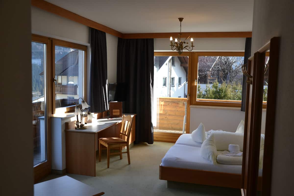 Une chambre d’hôtel équipée d’un lit, d’un bureau, d’une chaise et de grandes fenêtres donnant sur une maison et des arbres.