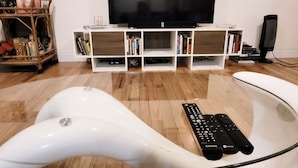 Una sala moderna con una televisión de pantalla plana sobre un estante blanco, una mesa de centro de vidrio y un control remoto sobre la mesa.