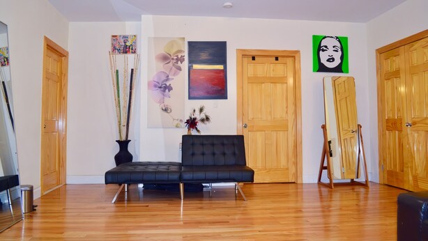 Una habitación con suelo de madera, un sofá negro, una puerta de madera y un cuadro en la pared.