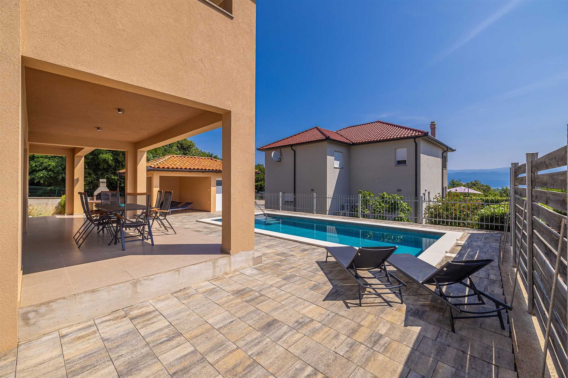 Zona de piscina con terraza cubierta, mobiliario exterior y vista a una casa con tejado de tejas rojas.