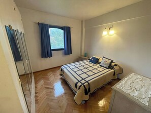 A bedroom with a bed, a nightstand, a window with curtains, and a wall-mounted light fixture.