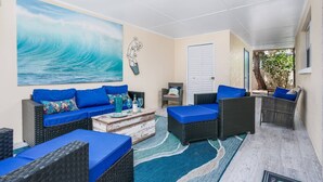 A sunlit room with a large wave painting, wicker furniture with blue cushions, a wooden coffee table, and a view of a tree outside.