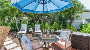 Un patio al aire libre con una sombrilla azul, sillas de mimbre, una mesa con sandía y copas de vino, y una valla de madera.