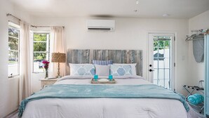 A bedroom with a bed, a bedside table with a lamp, a window with curtains, and an air conditioning unit.