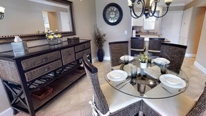 A dining area with a round glass table, wicker chairs, a dark wooden console table with baskets, a large mirror, and a wall clock. - White Sands & Ocean Waves Bld 5 2nd fl (Siesta Key)