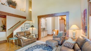 A spacious living room with a high ceiling, wooden beams, a staircase, a sofa set, a patterned rug, and a kitchen in the background. - Avon Duplex Between Beaver Creek & Vail Resorts! (Avon)