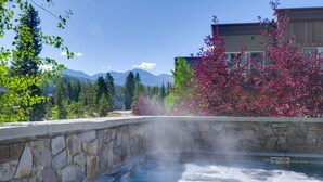 A hot tub with a stone wall, surrounded by trees and a building with a view of mountains. - Stunning Winter Park Condo Near Slopes! (Winter Park)