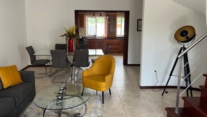 Un soggiorno moderno con un divano, una poltrona gialla e un tavolino da caffè in vetro. Si intravede una cucina sullo sfondo, con mobili a parete in legno e una finestra.