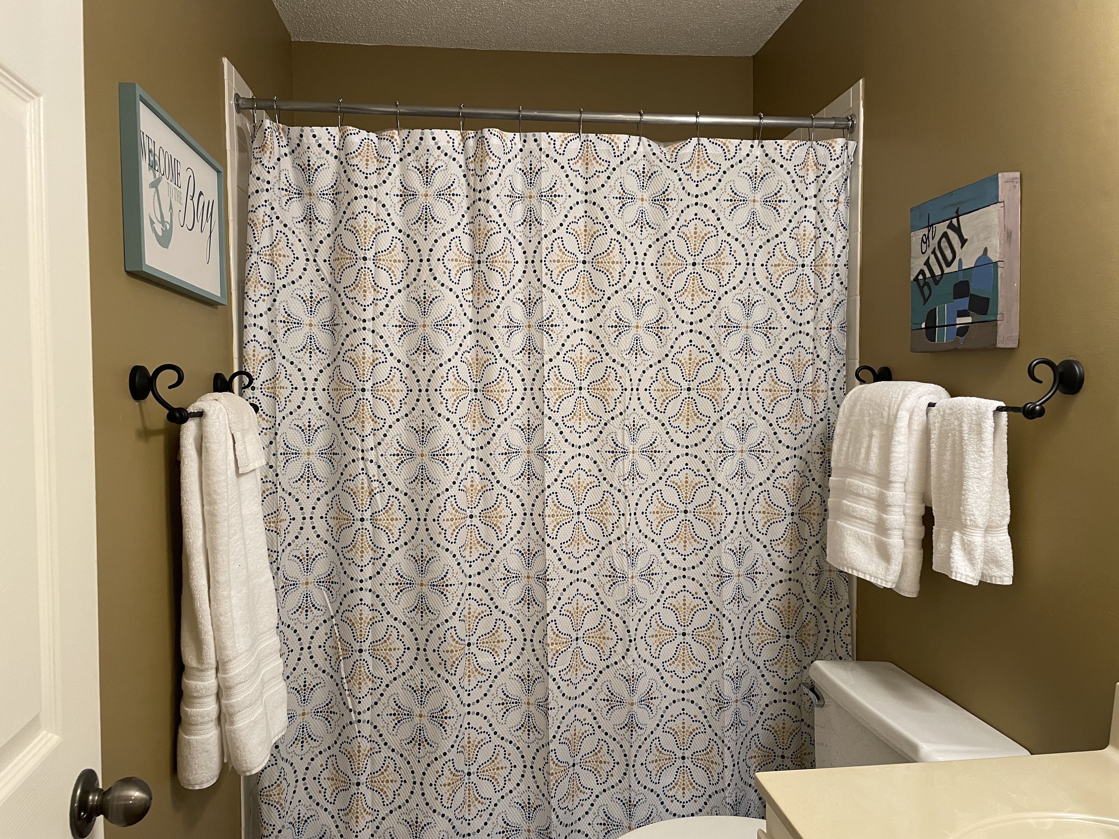 A bathroom with a patterned shower curtain, towels hanging on hooks, and a framed welcome sign.