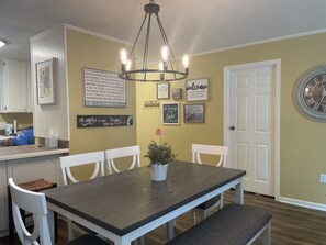 Un coin repas avec une table en bois, des chaises blanches et un lustre. Les murs sont décorés et il y a une horloge.