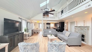 A spacious living area with a flat-screen TV, grey sofas, patterned armchairs, a wooden coffee table, and a kitchen in the background.