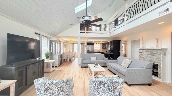 A spacious living area with a flat-screen TV, grey sofas, patterned armchairs, a wooden coffee table, and a kitchen in the background.