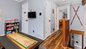 A room with a wooden floor, a table with a board game, a bookshelf with board games, a TV mounted on the wall, and a pair of skis hanging on the wall.