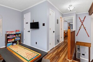 Una habitación con suelo de madera, una mesa con un juego de mesa, una estantería con juegos de mesa, un televisor montado en la pared y un par de esquíes colgados en la pared.