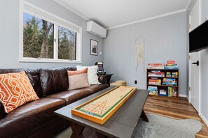 A living room with a brown leather sofa, a coffee table with a board game, a bookshelf with board games, and a window with a view of trees.