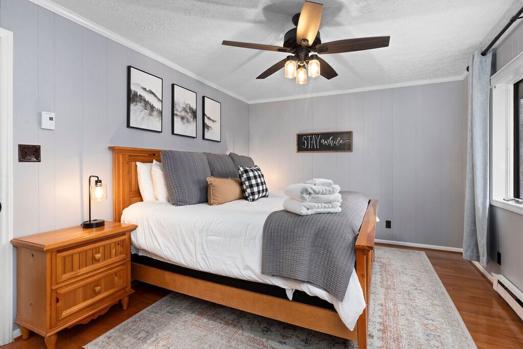 A bedroom with a bed, wooden nightstand, ceiling fan, wall art, and a motivational sign.