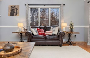 Ein gemütliches Wohnzimmer mit einem Ledersofa, zwei Beistelltischen mit Lampen, einer kleinen Weihnachtstanne und einem Bären-Motiv an der Wand.