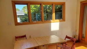 Una habitación con una mesa de madera y cuatro sillas, una ventana que da a árboles, y una puerta.