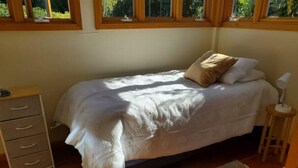 Una cama individual con una colcha blanca y dos almohadas, una mesita de noche de madera con una lámpara, una silla de mimbre y una alfombra tejida.