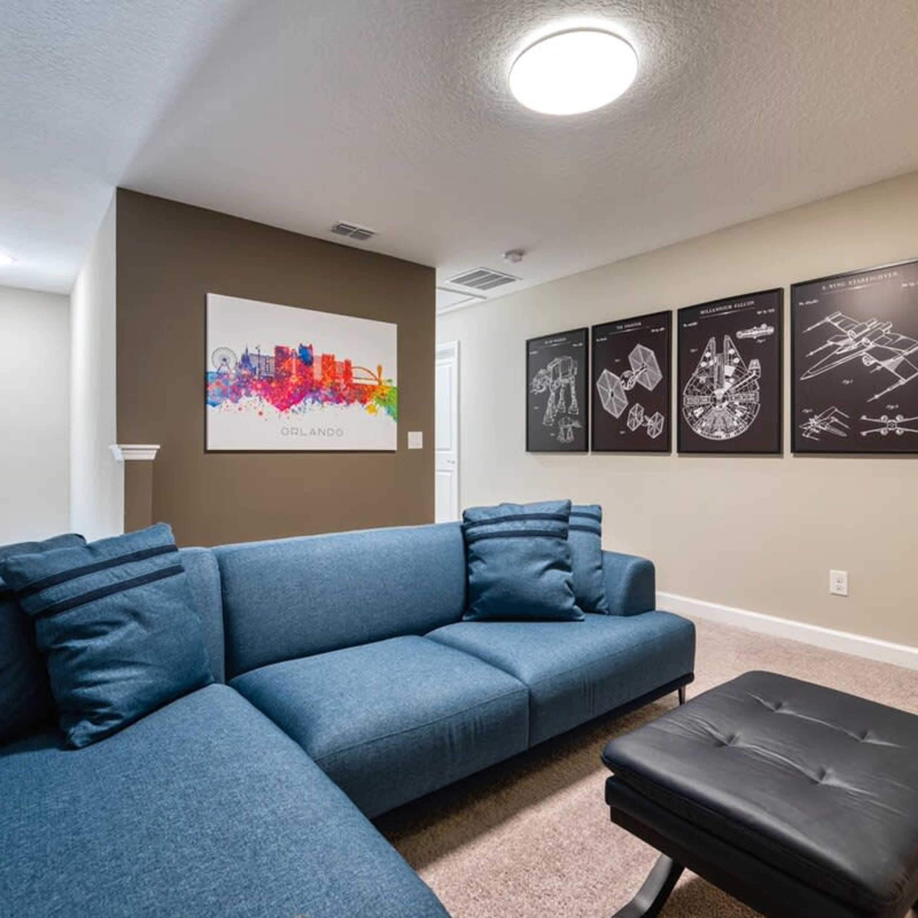 Un salon avec un canapé d’angle bleu, un pouf noir et une œuvre d’art représentant un paysage urbain accrochée au mur.