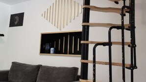 A modern living room with a grey sofa, a wooden bookshelf, and a black metal staircase.