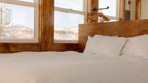 Un letto con lenzuola bianche in una stanza dalle pareti in legno e una finestra affacciata su un paesaggio innevato.