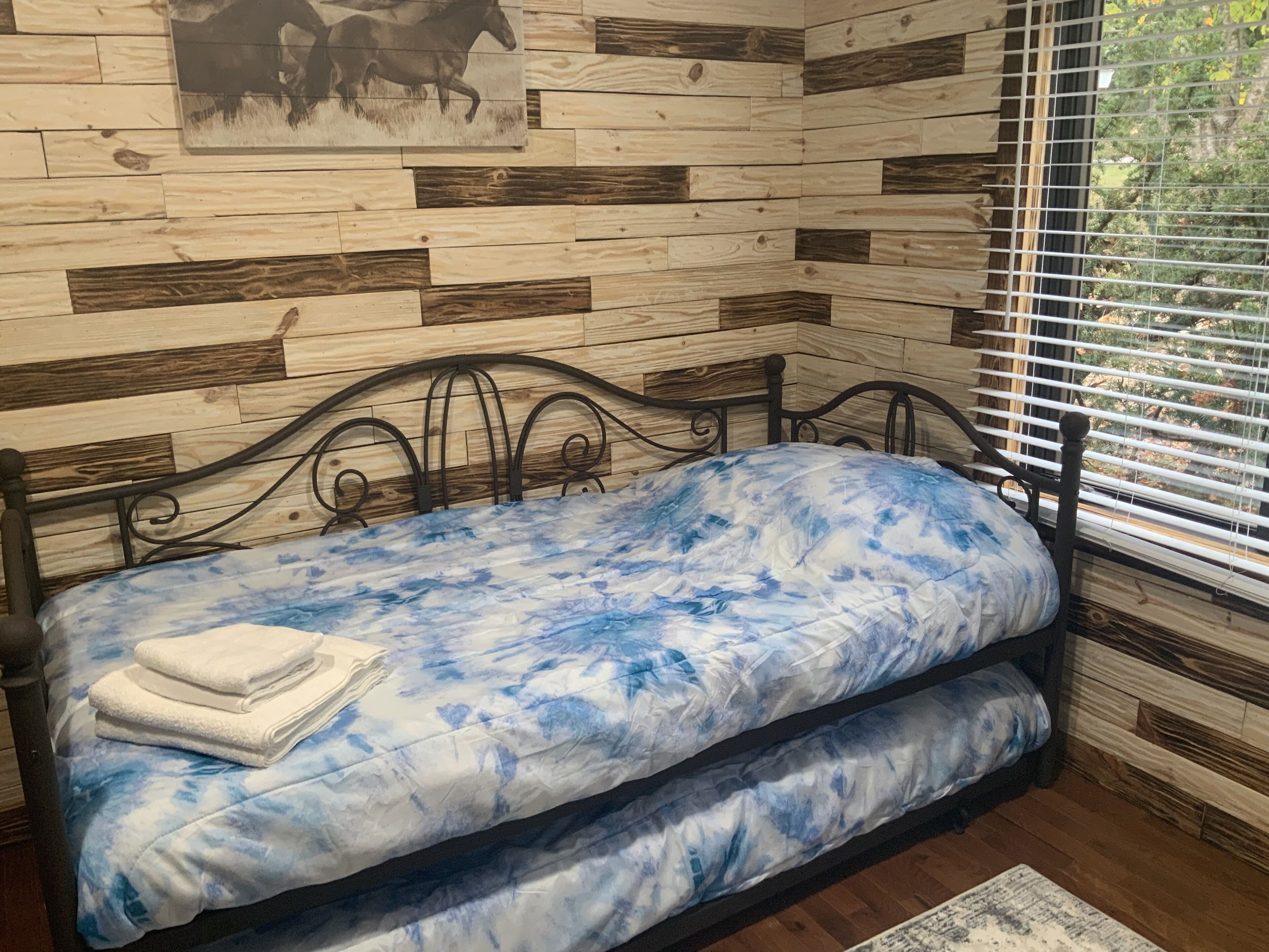 A bed with a blue and white patterned bedspread, a white pillow, and a folded white towel. The bed is positioned against a wall with a wooden plank design.