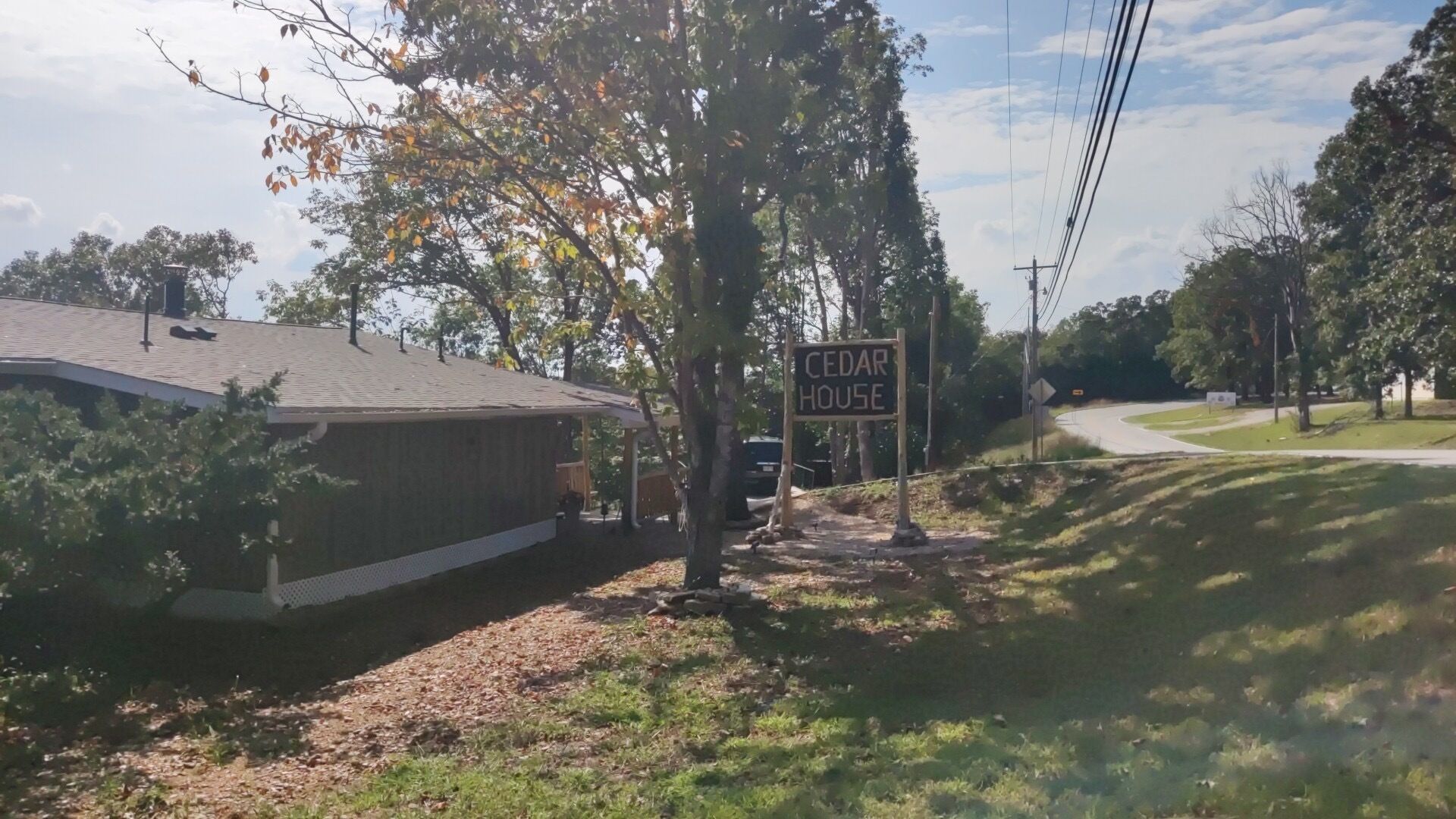 Una casa con un cartel que dice "CEDAR HOUSE" en un barrio residencial con árboles y una calle al fondo.