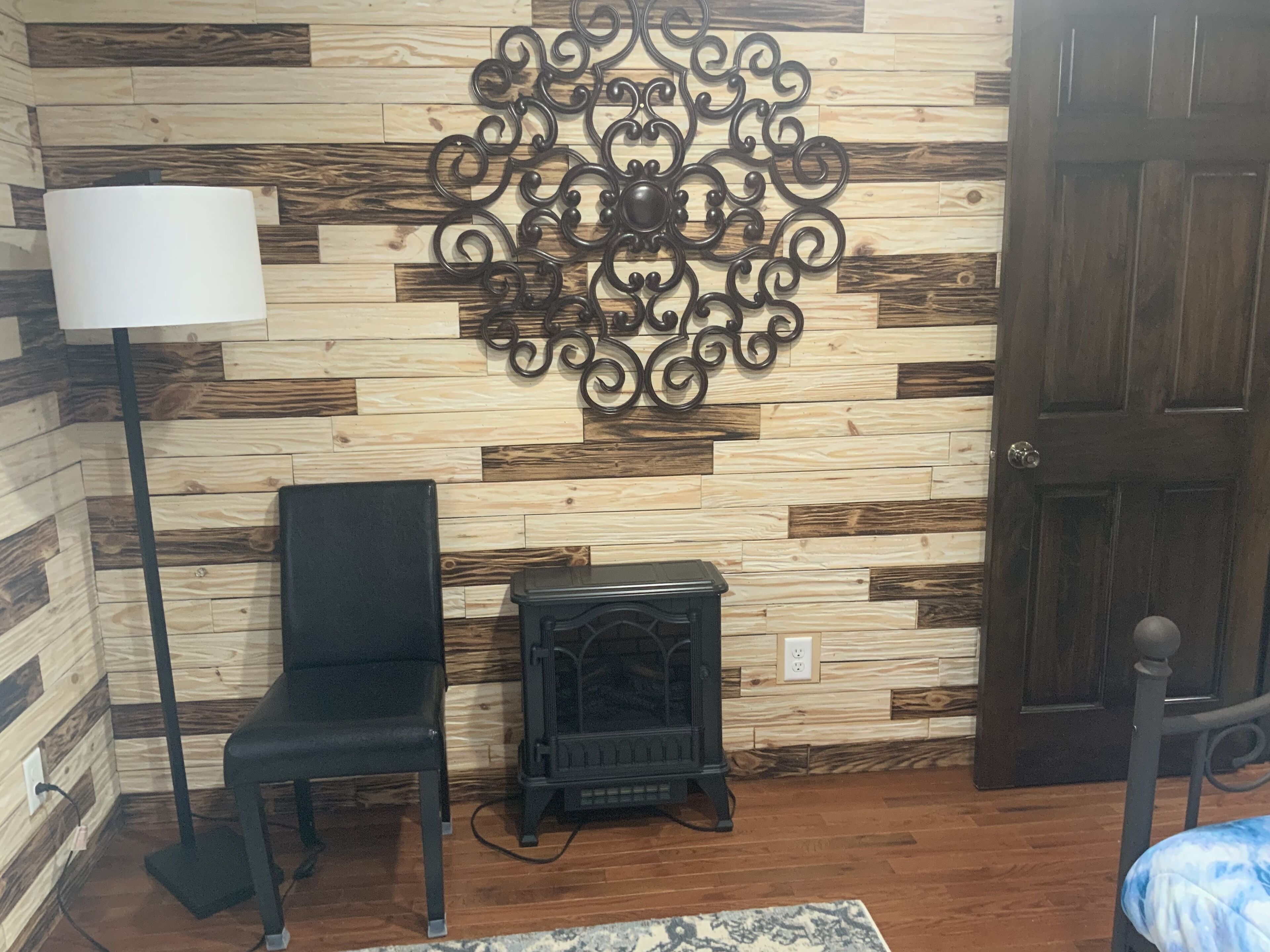 Une pièce avec un mur en bois, un élément décoratif en métal, une chaise noire, un radiateur noir, une lampe blanche et une porte.