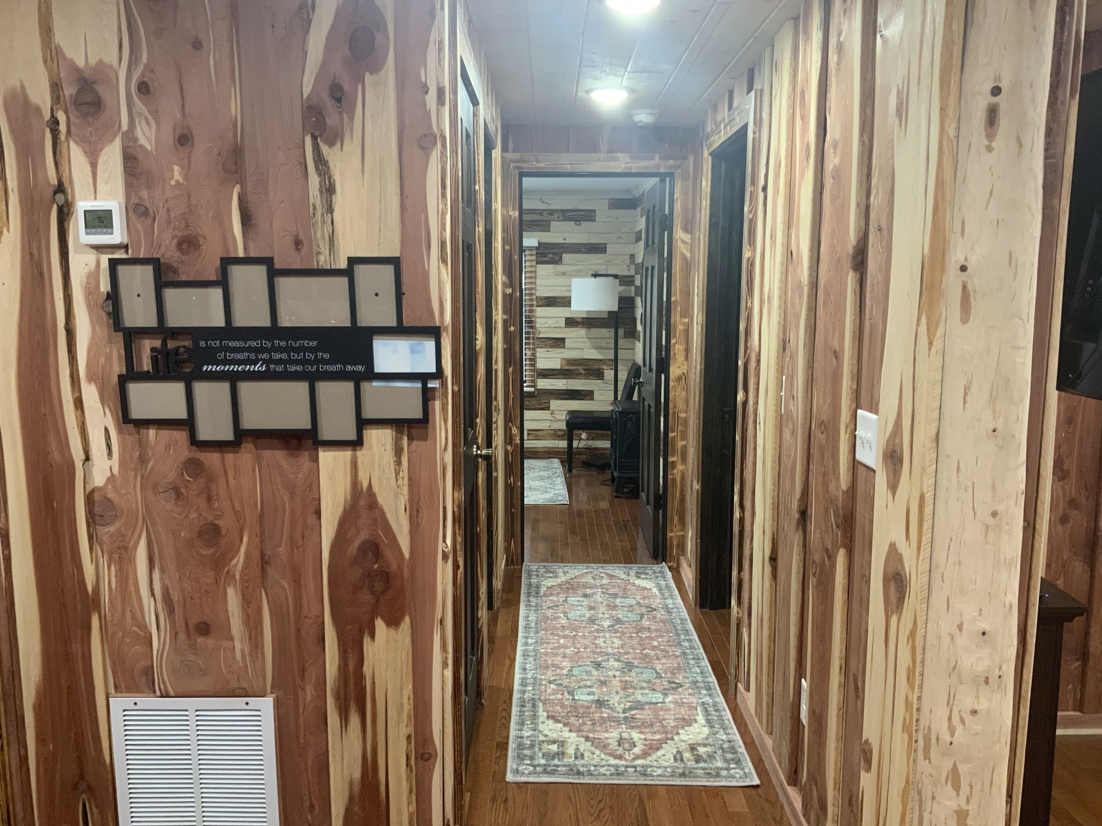 A wooden hallway with a patterned rug, a wall-mounted quote, and a thermostat.
