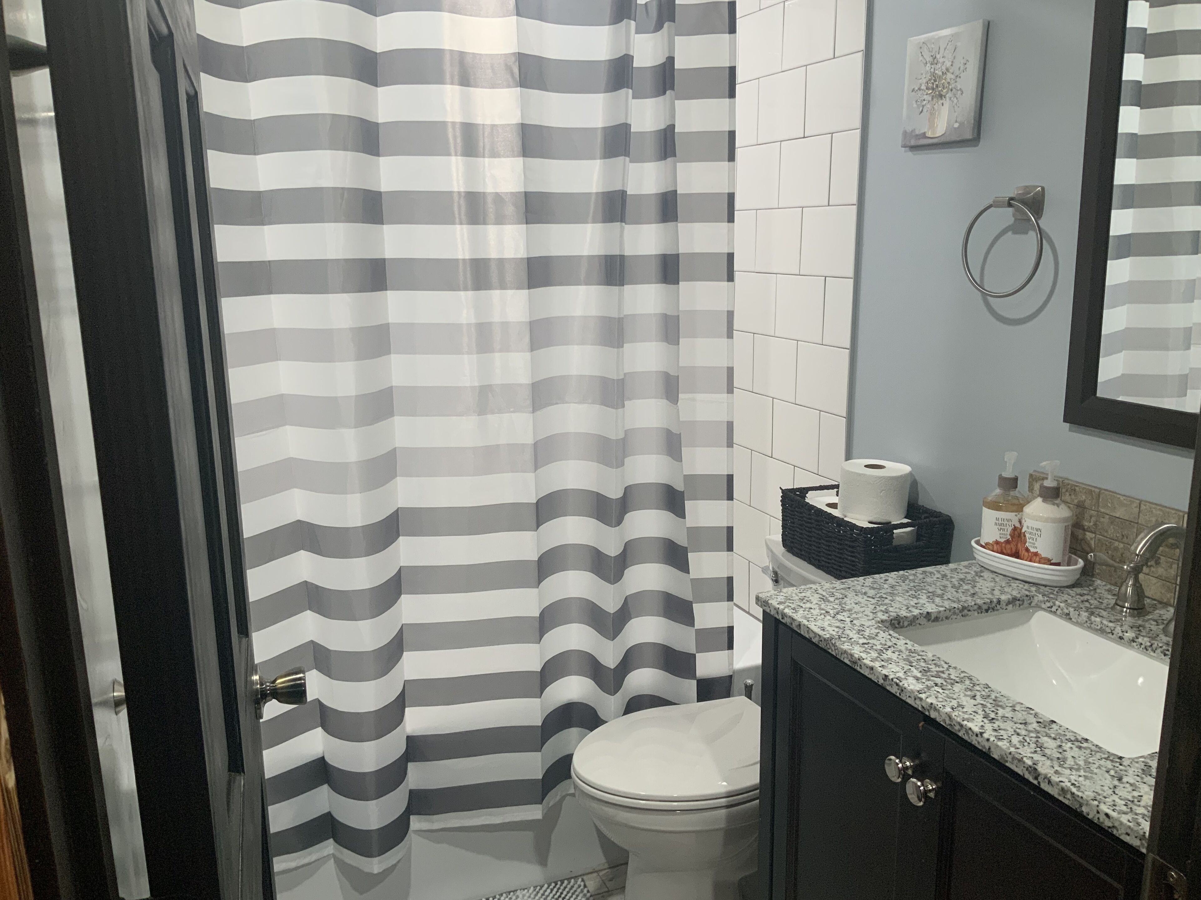 A bathroom with a striped shower curtain, white tiles, a toilet, a sink with a marble countertop, and a mirror.