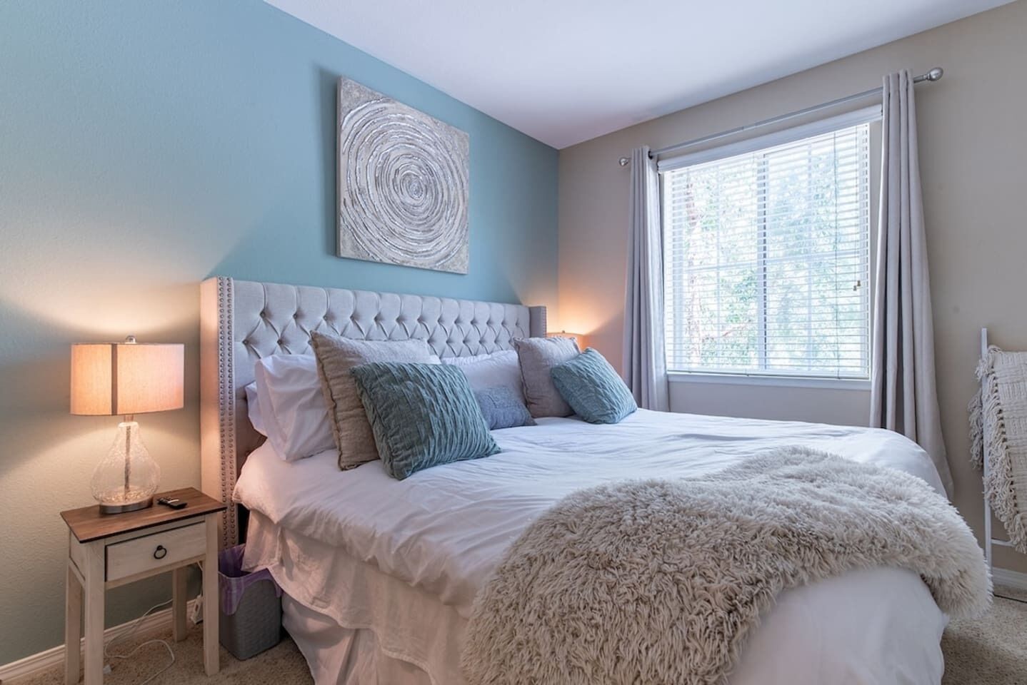 Een slaapkamer met een gestoffeerd hoofdeinde, een nachtkastje met een lamp, een groot raam met gordijnen en een kunstwerk aan de muur.