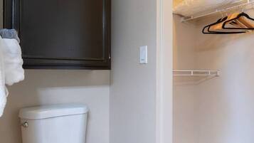 A bathroom with a toilet, a dark cabinet above it, and a walk-in closet with a hanging rack and a folded bed.