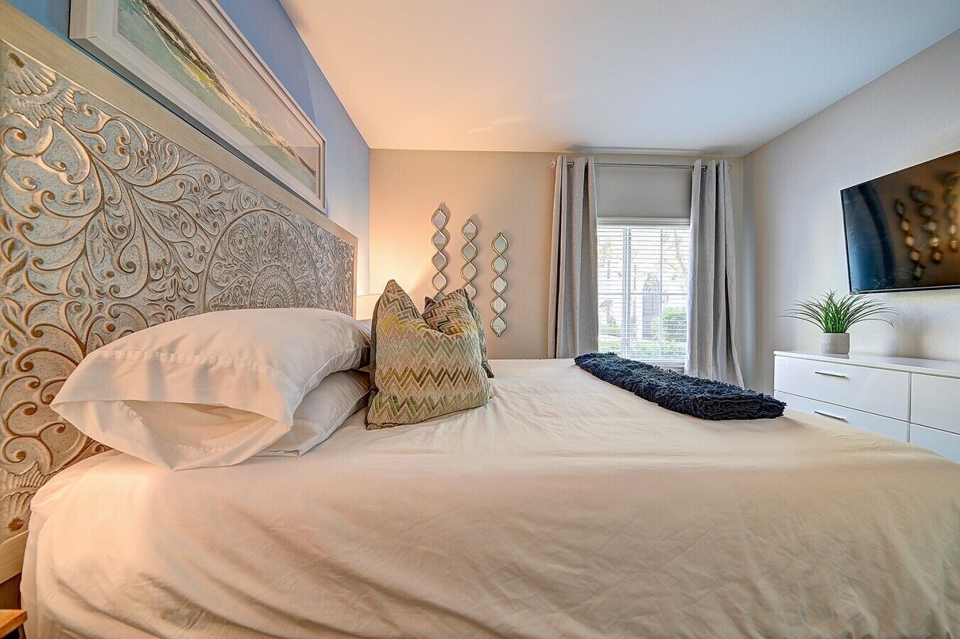 A bedroom with a large bed, a decorative headboard, a window with curtains, and a flat-screen TV on a white dresser.