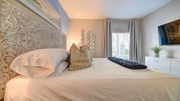 A bedroom with a large bed, a decorative headboard, a window with curtains, and a flat-screen TV on a white dresser.