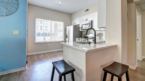 A modern kitchen with white cabinets, a stainless steel refrigerator, and a microwave. There are two stools and a window with blinds. - Sky Luxury 2BDRM Suite | Deluxe Living Cali (Valencia)