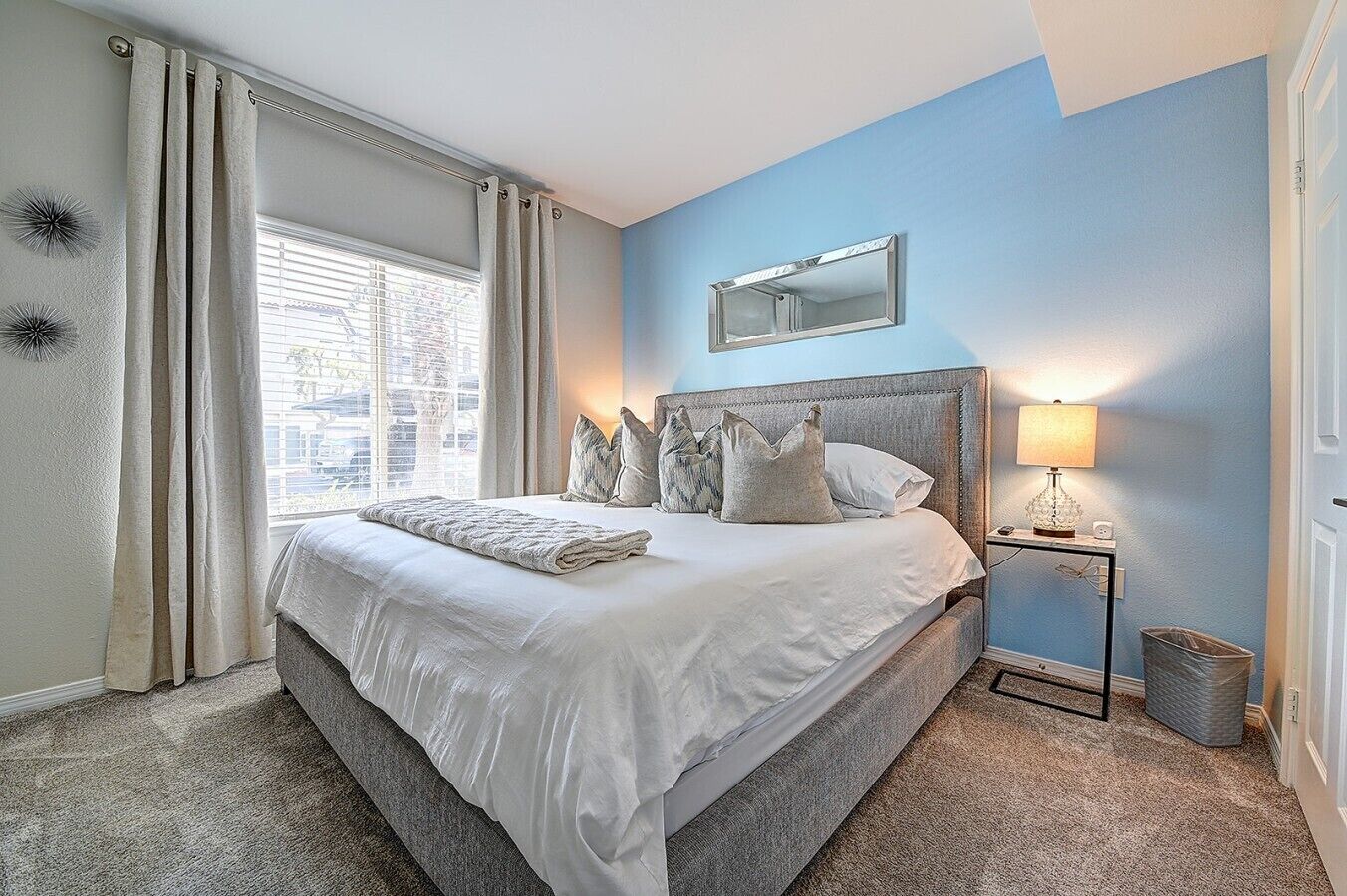A bedroom with a bed, a nightstand, a lamp, a mirror, and a window with curtains.